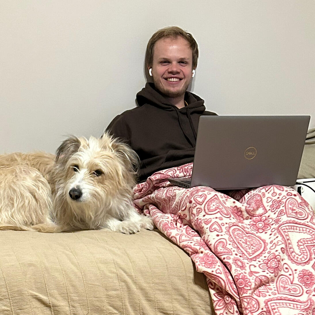 A man sitting on the bed with his laptop. He has earphones and blanket covering his legs. Big white dog is lying next to him.
