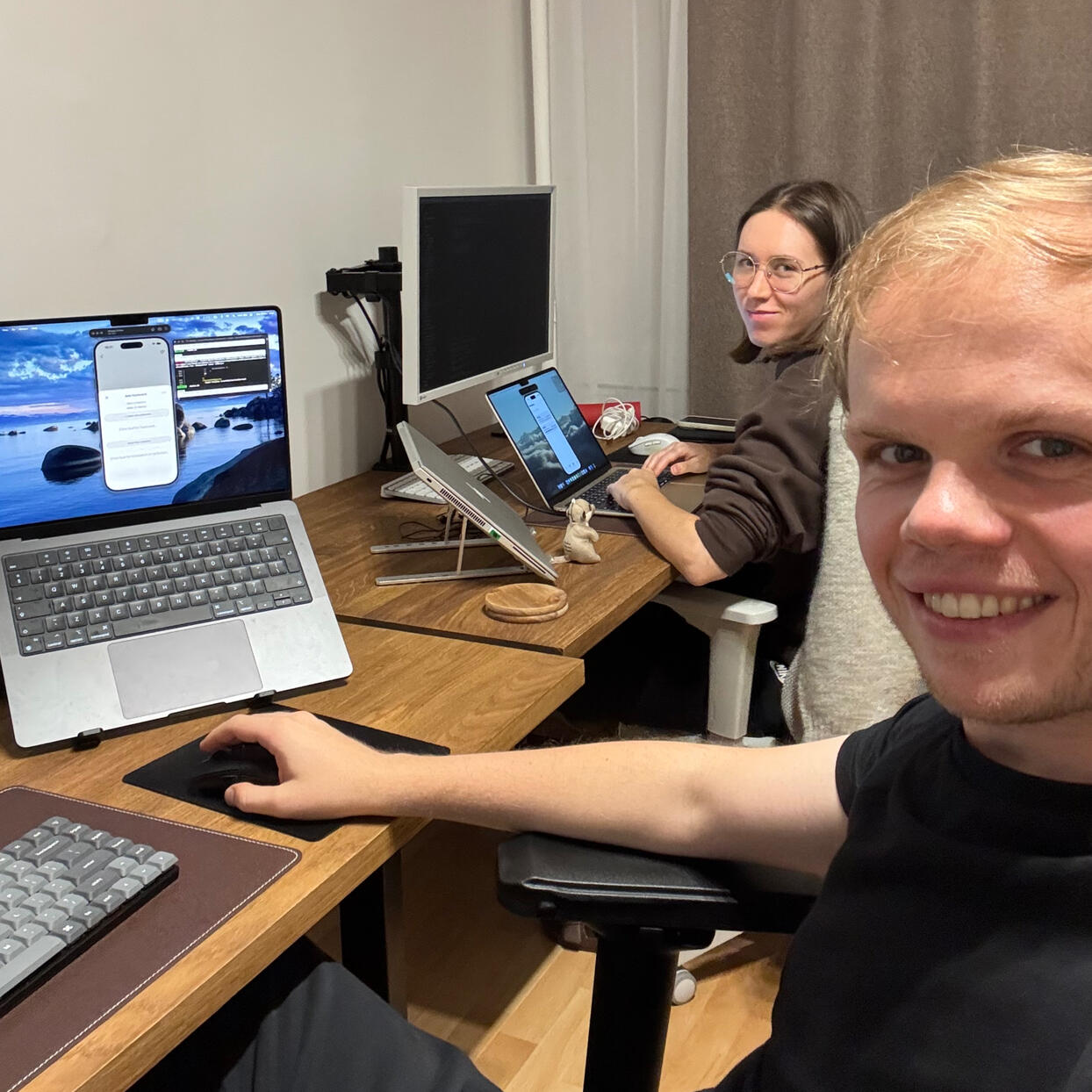 Foreground of the image shows a man sitting at the desk with his laptop on. In the background, a woman with glasses works on her laptop.