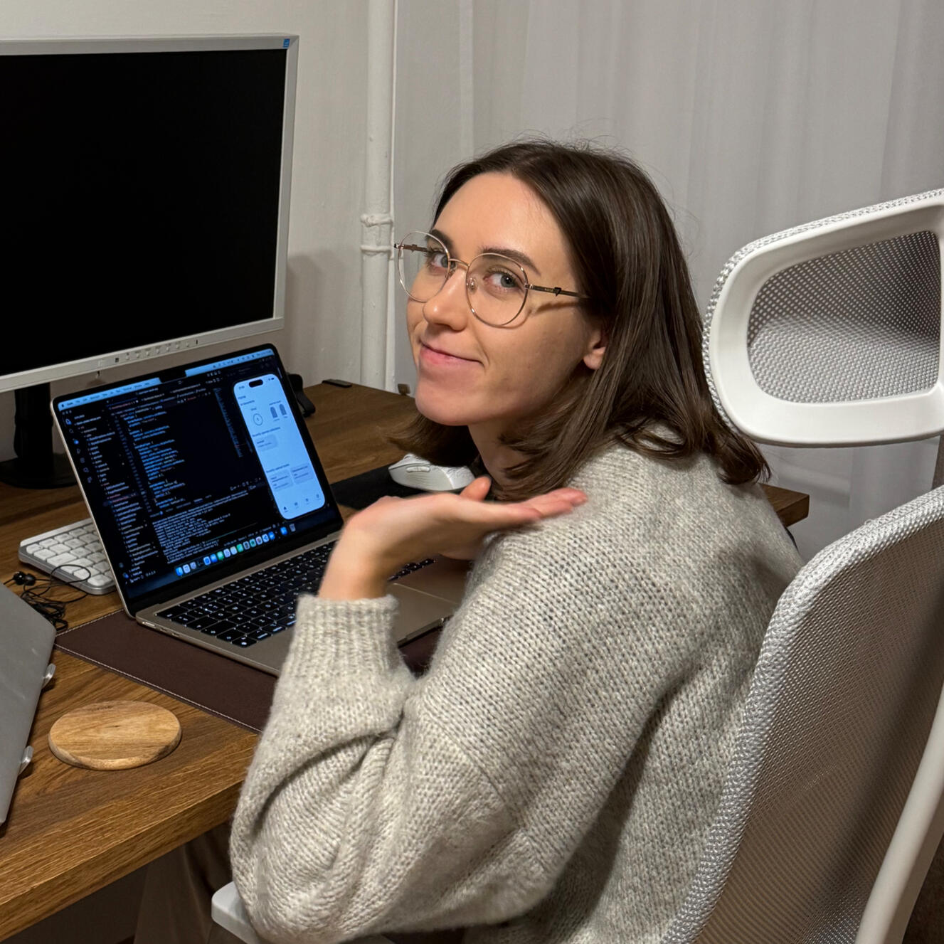 A woman with glasses sitting at the desk with her laptop up and running. There is mobile device emulator displayed on the laptop screen.